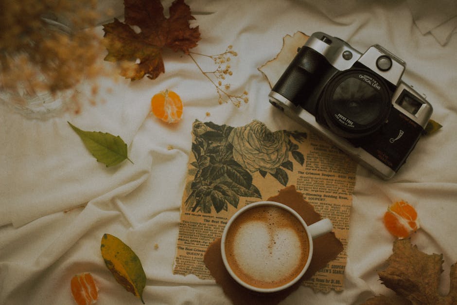 A cozy flat lay featuring a vintage camera, autumn leaves, a cup of frothy coffee, and newspapers.